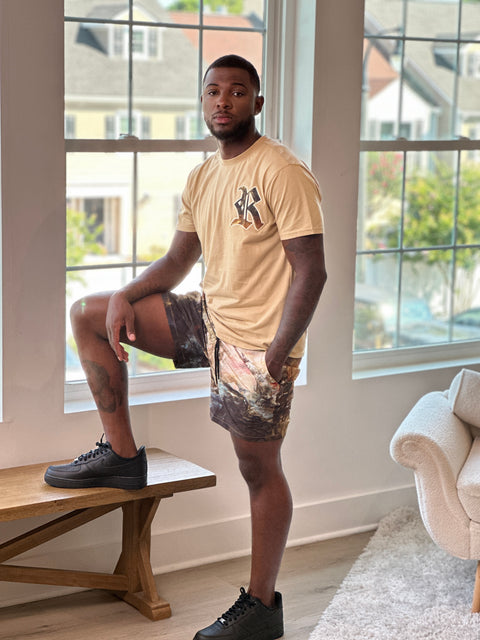 Man in beige t-shirt and patterned shorts standing in a room with large windows.