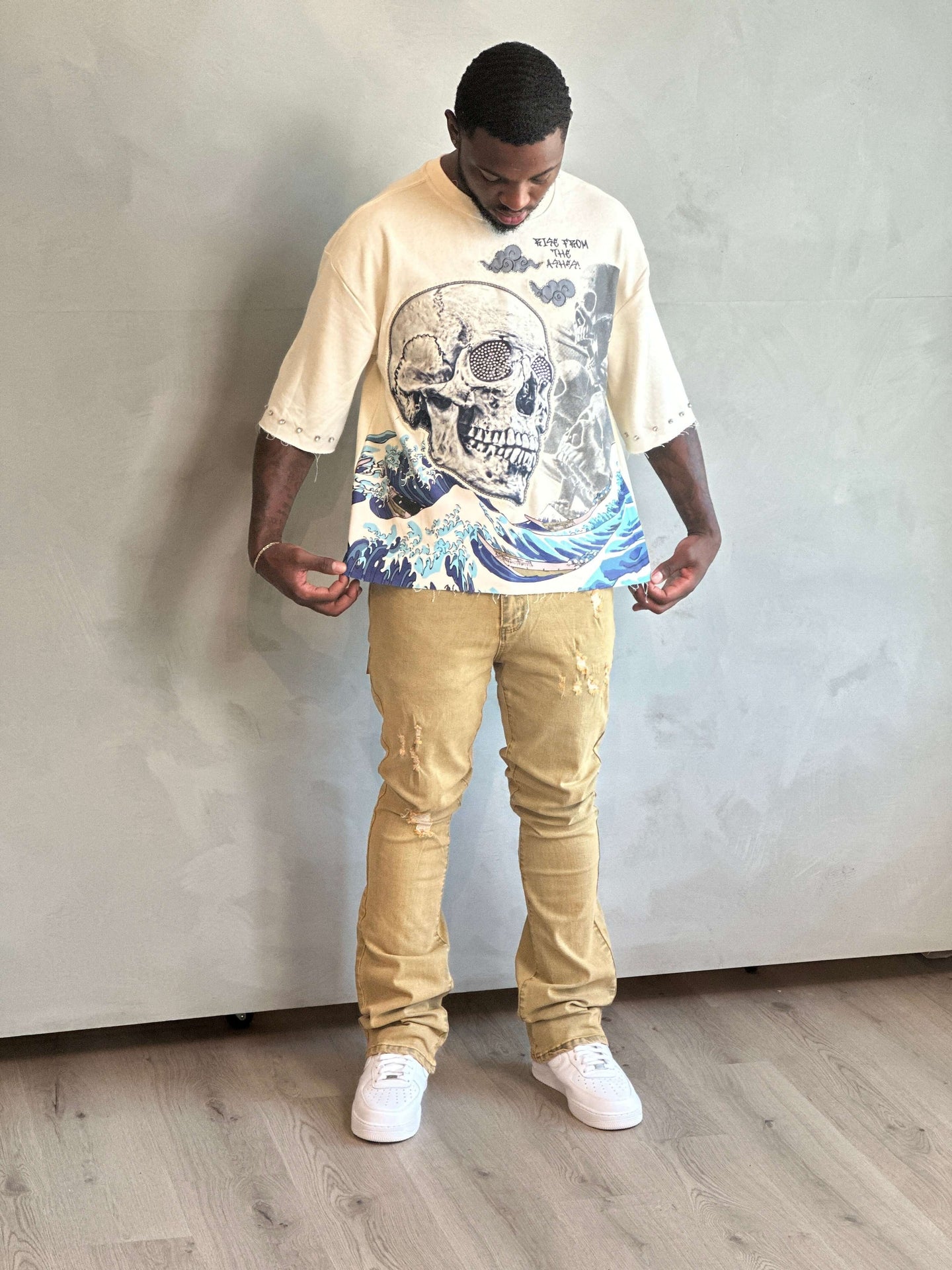 Man wearing a beige t-shirt with a graphic design and beige pants against a plain wall.