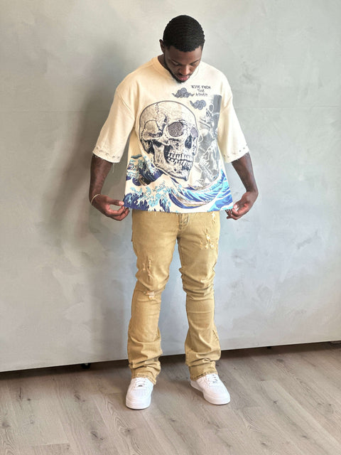 Man wearing a beige t-shirt with a graphic design and beige pants against a plain wall.