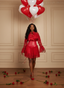 Woman in a red dress holding heart-shaped balloons against a beige wall with scattered roses on the floor.